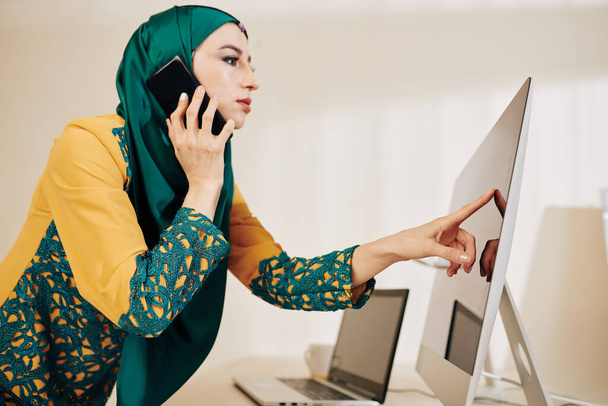 Mulher de negócios jovem franzina em hijab esmeralda bonita apontando para a tela do seu computador e falando ao telefone com o cliente ou parceiro de negócios
 - Foto, Imagem