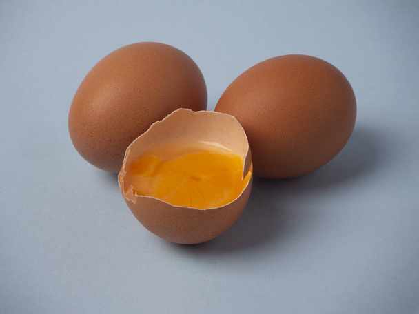 Kaksi raakaa ruskeaa kananmunaa ja rikkinäinen puolikas muna keltaisella keltuaisella lähikuva sinisellä pohjalla
.  - Valokuva, kuva