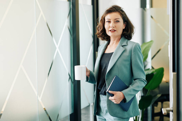 Porträt einer seriösen Geschäftsfrau mit einer Tasse Kaffee und einem Planer, der in die Kamera blickt - Foto, Bild
