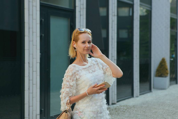 Young blond european woman using a smartphone while standing in the shopping district of a city, with house windows in the background. wearing sunglasses and handbag. - Foto, afbeelding