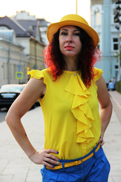 женщина с рыжими кудрявыми волосами, желтая шелковая блузка и желтая шляпа. Портрет по талии в городской среде
 - Фото, изображение