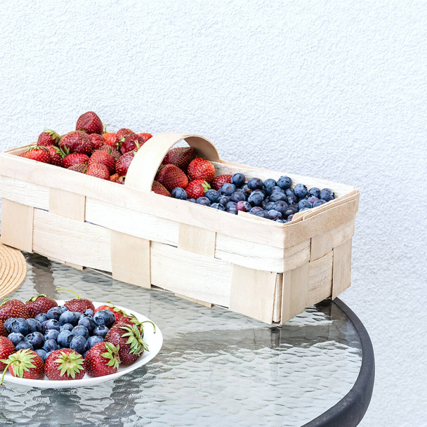 新鮮なイチゴとブルーベリーは、白い壁の漆喰を背景にガラスのテーブルの上のプレートとバスケットにあります。食品の概念、フラットレイアウト、園芸、夏、健康的な食事 - 写真・画像