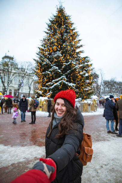 Следуйте за мной концептуальной женщиной, держащей за руку рождественскую елку на заднем плане на центральной городской площади. зимние путешествия
 - Фото, изображение