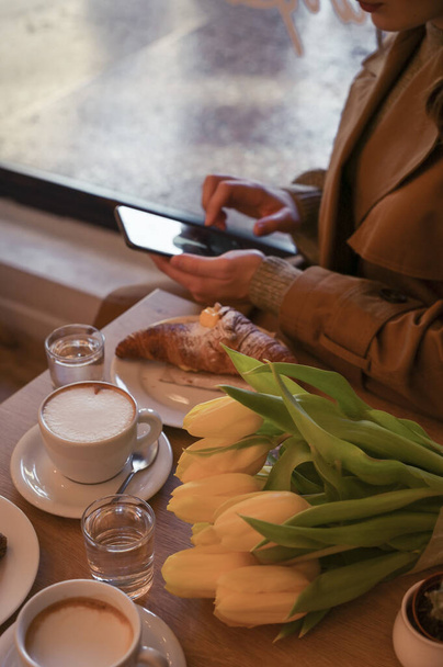 A young girl with a phone in a cafe, on the table is a bouquet of tulips and coffee with buns. Copy space - Foto, afbeelding