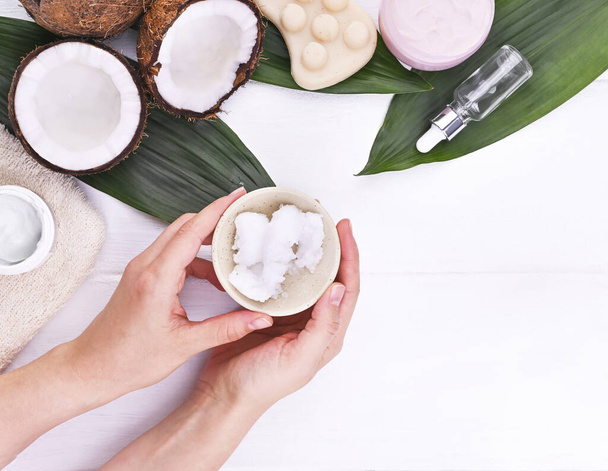Female hands hold coconut oil. The concept of natural cosmetics for beauty. Ripe coconuts and palm leaves on a white background. Copy space. Flat lay - Фото, изображение