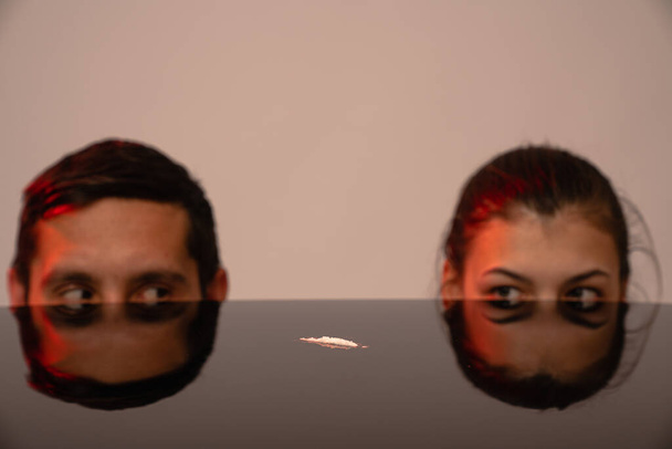 Line of cocaine lies in the middle of the black table, young man and girl peek out from the table, grey wall on the background - Foto, afbeelding