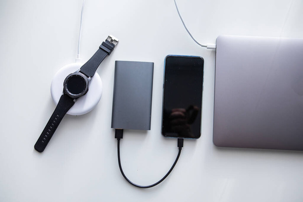 ноутбук з телефонною мишкою та смарт-годинник, що заряджається на бездротовому зарядному пристрої на білому столі
 - Фото, зображення