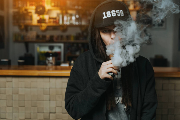 Egy fiatal csinos nő sapkában elszív egy elektromos cigarettát a gőzboltban. Hip-hop stílusban. Közelkép. - Fotó, kép