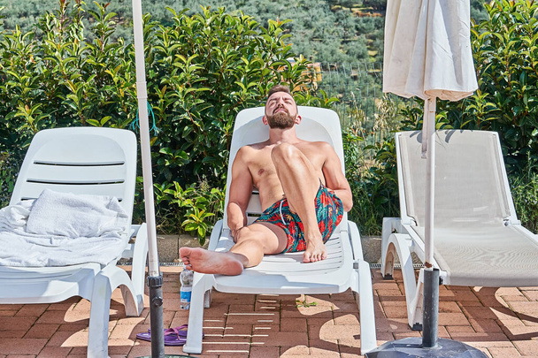 Hombre barbudo guapo tomando el sol en el sillón disfrutando con los ojos cerrados junto a la piscina durante el verano
 - Foto, Imagen