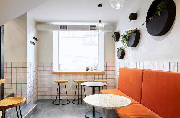 Stilvoll. Modernes Interieur des Cafés. Authentischer Stil mit Element des Dachgeschosses. Zwei runde schwarze und weiße Tische und orangefarbenes Sofa.Halbrunde Töpfe an der Wand mit Palmen. Weiße Fliesen mit orangefarbenen Leinen. Grüne, orange, weiße und graue Farben - Foto, Bild