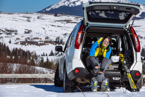 mujer cambiando botas para esquiar sentado en el maletero del coche. día soleado. viaje de invierno por carretera - Foto, Imagen