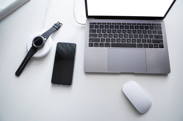 laptop με το ποντίκι του υπολογιστή του τηλεφώνου και έξυπνο ρολόι φόρτισης σε ασύρματο φορτιστή σε λευκό τραπέζι - Φωτογραφία, εικόνα