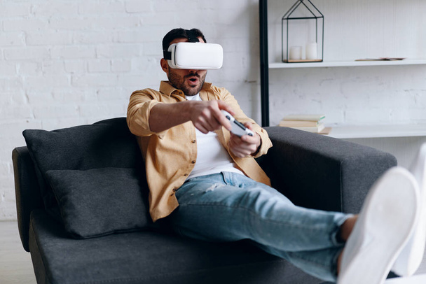 Joven hombre sonriente en auriculares VR jugando videojuego con joystick, acostado en el sofá
 - Foto, Imagen