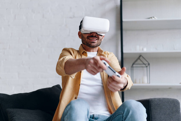 Jovem sorridente em óculos VR jogando videogame com joystick, deitado no sofá
 - Foto, Imagem