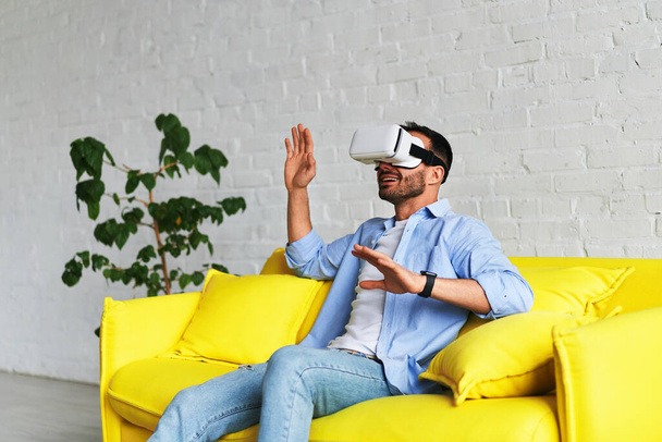 Front view of young smiling man using virtual reality glasses and showing his hand up - Valokuva, kuva