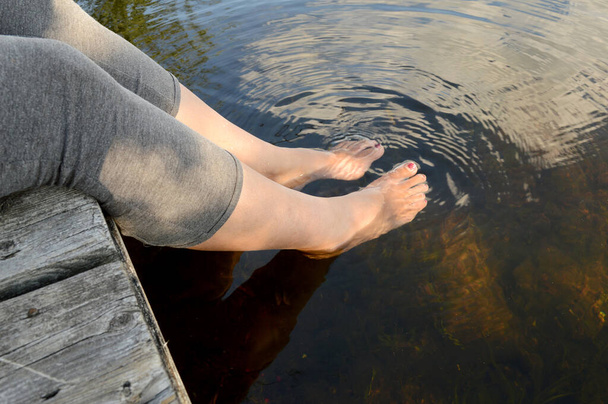 A woman splashes her feet in the cool river water as she rests on the dock during the heat of the summer. - Foto, Imagem