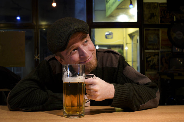 Rothaariger, fast irischer Typ sitzt in der Bar, trinkt ein Bier und blickt auf seine linke Seite - Foto, Bild