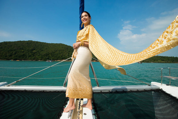 Asian Woman in Gold Yellow Thai Tradycyjny strój z biżuterią akcesoria uzyskać wiatry na luksusowy statek jacht w Green Ocean ładne niebo, Lady relaks na rejs noszenie starej sukienki formalnej - Zdjęcie, obraz