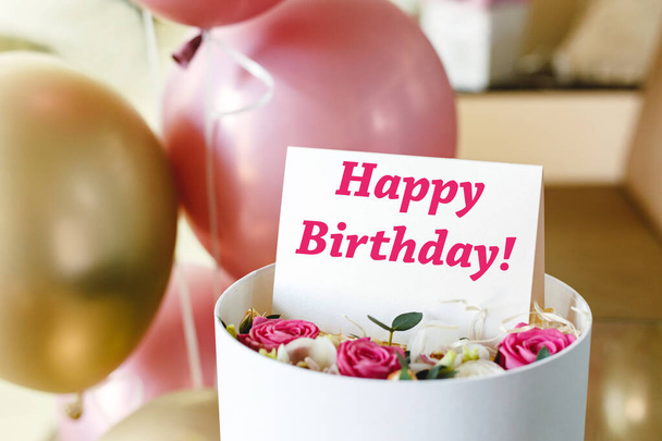 Happy Birthday text on gift card in flower box near festive pink and gold balloons. Beautiful bouquet of fresh flowers roses in box with greeting card Happy Birthday. - Foto, immagini