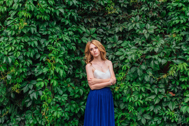 Portrait of a young beautiful woman with make up and curly blond hair, standing next to the wall of wild grape leaves. Copy space. - Photo, Image
