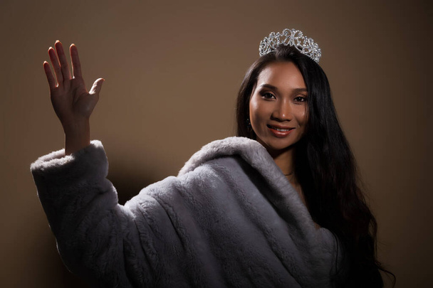 Portrait of Miss Pageant Beauty Contest in open back Evening fur Gown dress with sparkle light Diamond Crown, Asian Woman fashion make up black hair style, studio beige dark background dramatic - Φωτογραφία, εικόνα