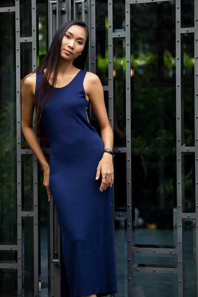 Beautiful Asian Long straight black hair tanned skin woman in dark blue open back dress slim, copy space, in front of glass door to look inside as peak and reflect her face - Valokuva, kuva
