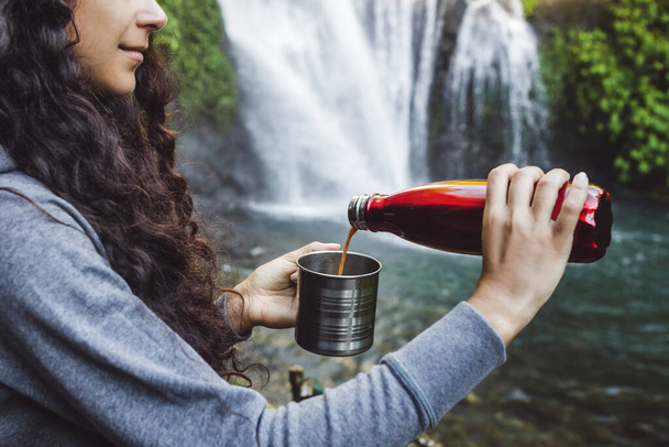 Vrouw handen gieten hete koffie uit rode thermosfles in stalen beker. Achtergrond van de tropische waterval. Geen afval eco concept. Wandeluitrusting. - Foto, afbeelding