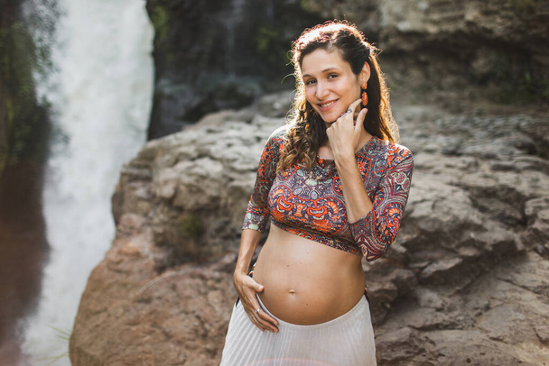 Молода вагітна жінка біля дивовижного водоспаду каскаду. Голий живіт і біла спідниця. Гармонія з природою. Tegenungan, Ubud, Indonesia - Фото, зображення