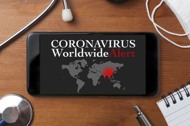 Coronavirus kavramı: steteskop, tıbbi maske ve dünya haritası olan diğer nesneler ve ekranda uyarı metni bulunan bir akıllı telefon - Fotoğraf, Görsel