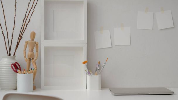 Detailní pohled na minimální designérské pracoviště s rámečky, malířskými nástroji, dekoracemi a notebookem na bílém stole s lepicí poznámkou na cihlové stěně  - Fotografie, Obrázek