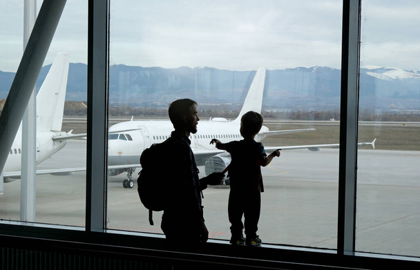 Silhouette eines Kindes und seines Vaters, die das Flugzeug durch das Fenster des Flughafenterminals betrachten. Lebensstil der Familie. - Foto, Bild