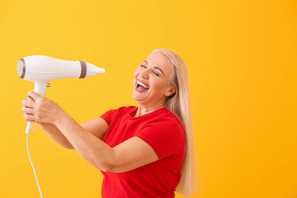 Beautiful mature woman with hair dryer on color background - Photo, Image