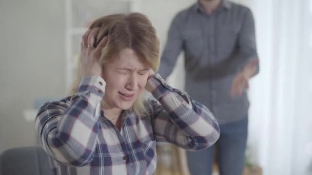 Close-up portrait of Caucasian woman closing ears with hands and shaking head as unrecognizable man yelling at the background. Depressed wife arguing with spouse indoors. Breakup, conflict, divorce. - Footage, Video