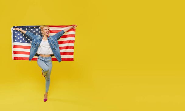 Une Américaine. Joyeux jeune femme en denim tenant drapeau des États-Unis isolé sur fond jaune
 - Photo, image
