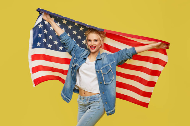 Americká holka. Šťastná mladá žena v džínových šatech drží vlajku USA izolované na žlutém pozadí - Fotografie, Obrázek
