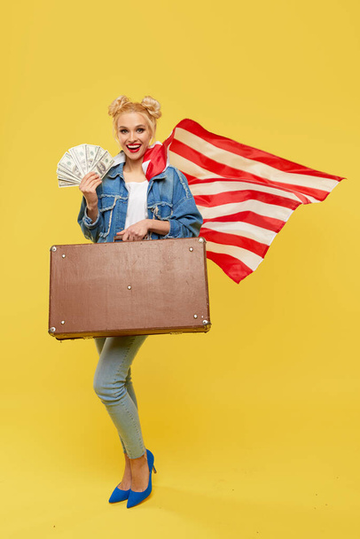 Una joven con una bandera americana lleva en sus manos una maleta de viaje y billetes de dinero. El concepto de ganar la lotería. Rostro alegre sorprendido
. - Foto, Imagen