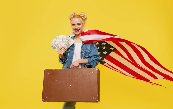 Elinde Amerikan bayrağı olan genç bir kadın elinde bir seyahat çantası ve para banknotları tutuyor. Piyango kazanma fikri. Şaşırmış neşeli yüz. - Fotoğraf, Görsel