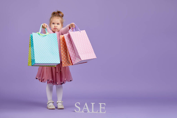 Sale of childrens products. Portrait of fun little child girl  on purple background holding shopping bags, package. Looking at the camera. Banner. Space for text.  - Photo, Image
