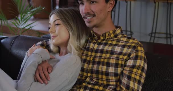 Front view of a mixed race man and a Caucasian woman enjoying time at home together, sitting on a couch in their sitting room, watching television and talking, embracing. - Video, Çekim
