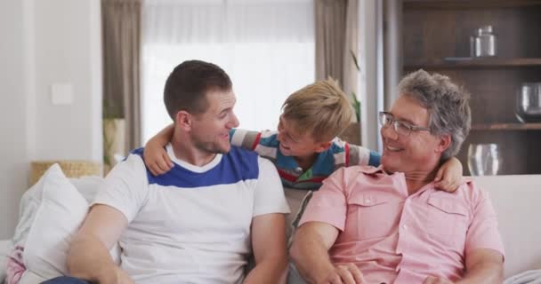 Front view close up of a Caucasian man enjoying his time in an apartment, sitting on a couch with his son and father, talking, looking at camera, embracing, in slow motion - Video