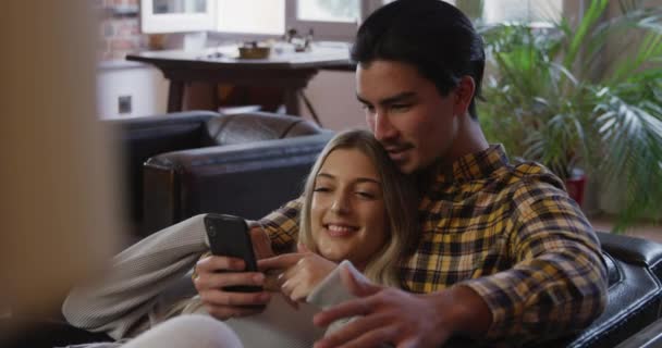 Front view of a mixed race man and a Caucasian woman enjoying time at home together, sitting on a couch in their sitting room, talking, embracing, the woman using smartphone. - Video, Çekim