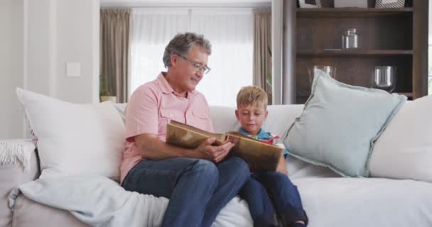Front view of a senior Caucasian man enjoying his time in an apartment, sitting on a couch with his grandson, looking at family photos, in slow motion - Video, Çekim