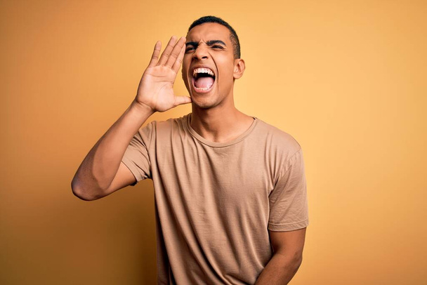 Nuori komea afrikkalainen amerikkalainen mies päällään rento t-paita seisoo yli keltainen tausta huutaa ja huutaa kovaa puolelta käsin suulla. Viestintäkäsite
. - Valokuva, kuva