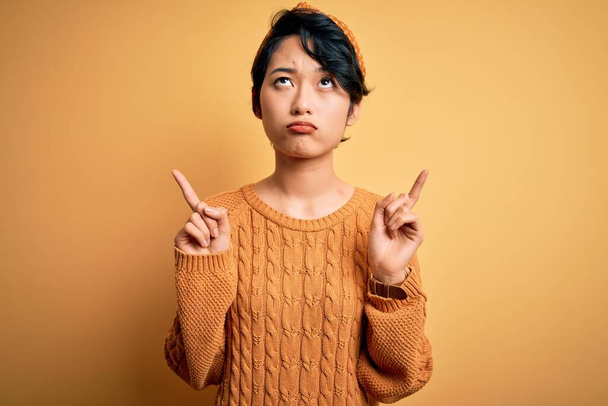 Fiatal gyönyörű ázsiai lány visel alkalmi pulóver és diadem áll a sárga háttér felfelé mutató néz szomorú és ideges, jelezve irányt ujjak, boldogtalan és depressziós. - Fotó, kép