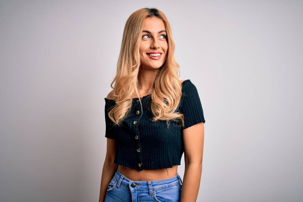 Young beautiful blonde woman wearing casual t-shirt standing over isolated white background looking away to side with smile on face, natural expression. Laughing confident. - Fotó, kép