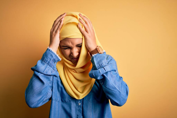 Mladá krásná žena s kudrnatými vlasy nosí arabsky tradiční hidžáb přes žluté pozadí trpí bolestí hlavy zoufalé a vystresované, protože bolest a migréna. Ruce za hlavu. - Fotografie, Obrázek