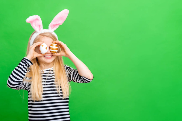 Girl in rabbit bunny ears on head on green studio background. Cheerful crazy smiling happy child. Easter painting eggs on eyes. Easter and holidays. Copy space - 写真・画像