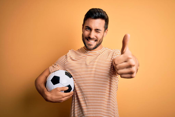 黄色の背景の上にサッカーボールを保持しているサッカーを再生ひげを持つハンサムな選手の男は、手で積極的なジェスチャーを行うことを承認し、親指を笑顔と成功のために幸せ。勝者のジェスチャー. - 写真・画像