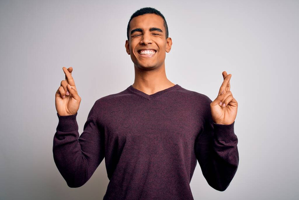 Junger gutaussehender afrikanisch-amerikanischer Mann in lässigem Pullover vor weißem Hintergrund gestikulierend, lächelnd mit Hoffnung und geschlossenen Augen. Glück und abergläubisches Konzept. - Foto, Bild