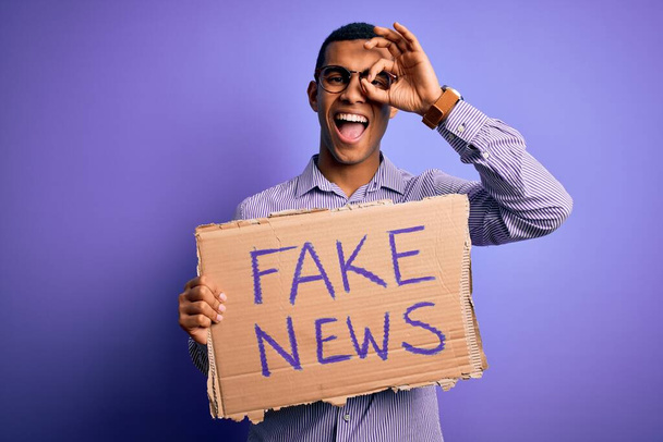 Jonge knappe Afro-Amerikaanse man protesteert met spandoek met nep nieuwsbericht met vrolijk gezicht lachend doen ok teken met hand op oog kijkend door vingers - Foto, afbeelding